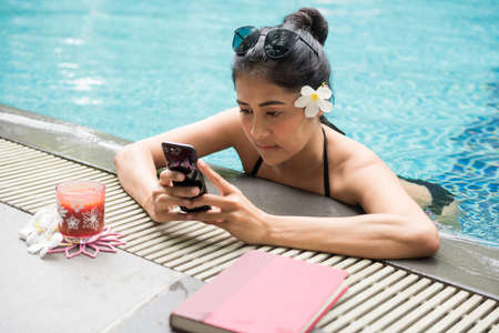 Attractive sexy young Asian woman with black two piece swimsuit in swimming pool play social media and chat by phone. Beautiful girl relax during summer vacation. Holiday maker in tropical country.の写真素材