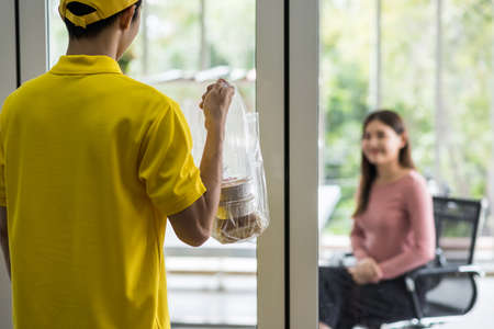 Delivery man in yellow uniform deliver cake dessert in plastic bags to blurred businesswoman customer in office meeting room. Happy girl wait for food. Lifestyle in working day. New normal conceptの写真素材