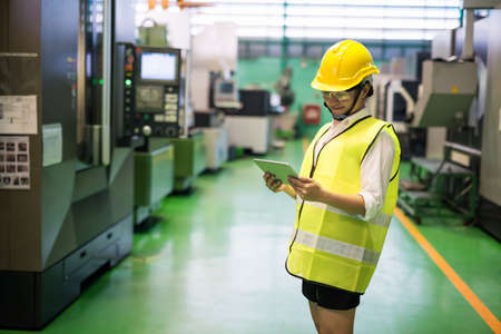 Waist up Asian smiling worker in hardhat and protection glasses using tablet to check transistor microchip machines in factory workshop with copy space. Manufacturing industry with technology.の写真素材