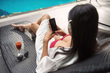 sexy Asian beautiful woman in red swimsuit play social media, chat, shoping online by mobile phone at bed chair of swiming pool. Female relax in summer at luxury hotel resort.の写真素材
