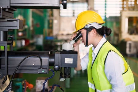 Waist up female worker with hardhat look at microscope to check quality control of glass productivity in manufacturing factory. Manufacture industry to produce with Quality assurance QA or QC.の写真素材