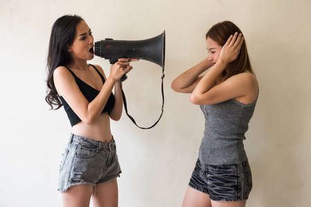 Bossy Asian woman shouting by megaphone while her friend close eyes to avoid loudly noise.の写真素材