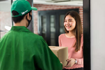 Smiling Asian woman receive delivery parcel from courier man with face mask at door inside apartment or condo. New normal to order goods online by application during  omicron pandemic.の写真素材