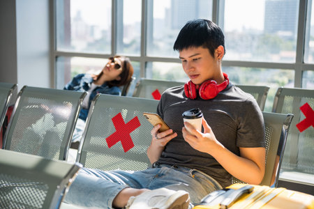Young Asian man using smartphone and drink coffee on waiting area with social distancing sign at...