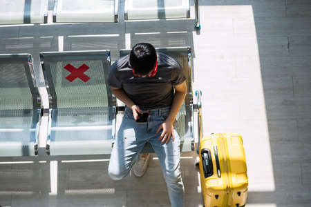 top view of Asian young man using phone at airport departure waiting seat with yellow suitcase.の写真素材