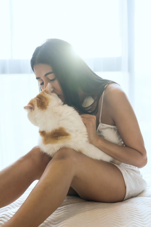 Beautiful sexy Asian woman in pajama hold and kiss her yellow Exotic shorthair cat on bed with...