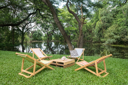 Relax wooden deckchair and table on green grass yard by pond with beautiful big trees and reflection on water. Outdoor cafe and restaurant.の写真素材
