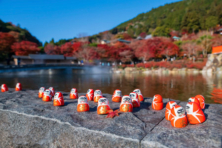 Daruma dolls with maple leaf by pond in autumn of Katsuo-ji temple, Minoh, Osaka, Japan.  Famous...