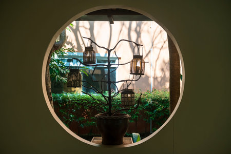 lanterns hanging on tree branch in flower pot decor by circle window with green garden. Home design...