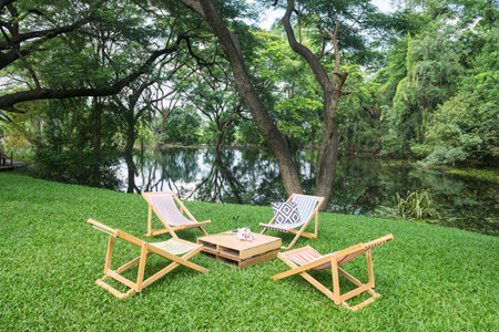 Wooden bedchair and table on green grass garden by pond with beautiful big trees and reflection on water. Outdoor cafe and restaurant.の写真素材