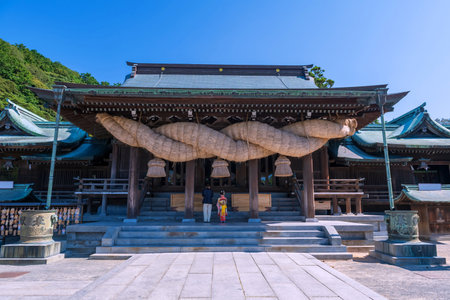 Cute girl in kimono sacred Miyajidake Shrine, largest shimenawa sacred ropes hangs across entrance to main shrine, against blue sky, Fukutsu, Fukuoka, Kyushu, Japan. Famous travel destination.の写真素材