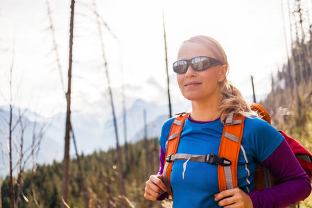 Woman hiking on trail in sunshine forest and Tatra Mountains in Poland. Recreation and healthy lifestyle outdoors in nature. Beauty blond hiker with backpack looking at camera.の写真素材