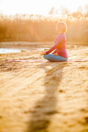 Young woman doing yoga near lake outdoors, meditation. Sport fitness and exercising in nature, autumn sunsetの写真素材