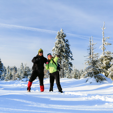 Happy man and woman hikers trekking on snow trail in winter mountains. Young couple on white and cold vacations in winter forest, beautiful natural landscape.の写真素材