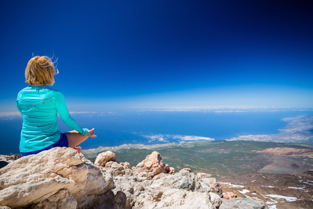 Young woman doing yoga meditation outside natural beautiful inspirational environment, fitness and exercising motivation and inspiration in sunny mountains over blue sky and ocean sea.の写真素材