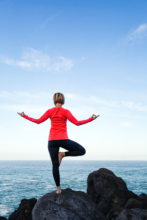 Woman meditating in yoga vrksasana tree pose silhouette at the ocean, beach and rock mountains. Motivation and inspirational fit and exercising. Healthy lifestyle outdoors in nature, fitness concept.の写真素材
