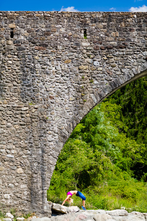 Woman meditating in yoga pose under a medieval stone bridge near river in beautiful mountains. Motivation and inspirational stay fit and exercise. Healthy lifestyle outdoors in nature, fitness concept.の写真素材