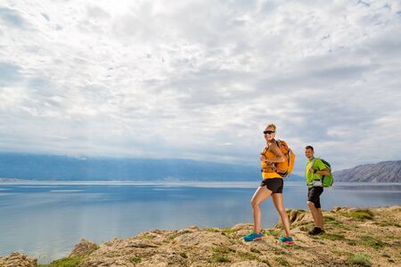 Happy couple hikers trekking in summer mountains on island seaside. Young woman and man walking with backpack on rocky mountain trail. Camping and looking at beautiful inspirational landscape view.の写真素材