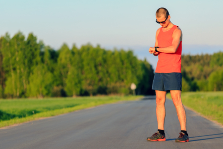 Man runner running on country road in summer sunset. Checking sports watch, ready to run. Young road runner athlete male training and doing workout outdoors in nature.Sport training and fitness concept outdoors.の写真素材
