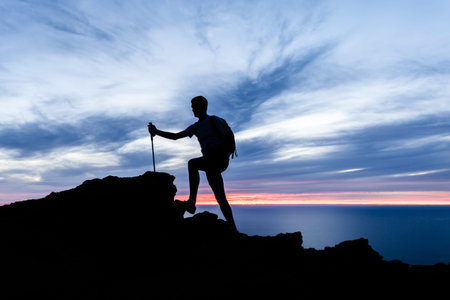 Man hiking silhouette in mountains, sunset and ocean inspirational landscape. Male hiker with walking sticks on top of mountain looking at beautiful night sea landscape.の写真素材