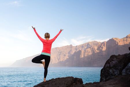 Woman meditating in yoga vrksasana tree pose silhouette at the ocean, beach and rock mountains. Motivation and inspirational fit and exercising. Healthy lifestyle outdoors in nature, fitness concept.の写真素材