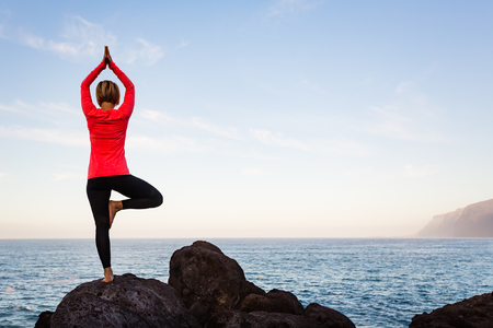Woman meditating in yoga vrksasana tree pose silhouette at the ocean, beach and rock mountains. Motivation and inspirational fit and exercising. Healthy lifestyle outdoors in nature, fitness concept.の写真素材