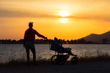 Silhouette of mother with child in stroller enjoying motherhood at sunset landscape. Walking or jogging woman with pram on a beach at sunset. Family relaxing at beautiful inspirational mountains landscape.の写真素材