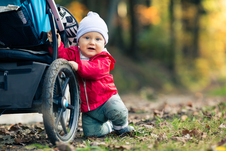 Baby boy playing in autumn forest with stroller, outdoors fun. Happy child on hiking trip in woods, looking at camera.の写真素材
