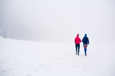 Girls running together on snow in winter mountains. Sport, fitness inspiration and motivation. Two women partners trail running in mountains, winter day. Female trail runner jogging outdoors.の写真素材