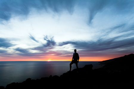 Celebrating or meditating man looking at sunset ocean. Hiker with backpack on top of mountain looking at beautiful inspirational night landscape.の写真素材