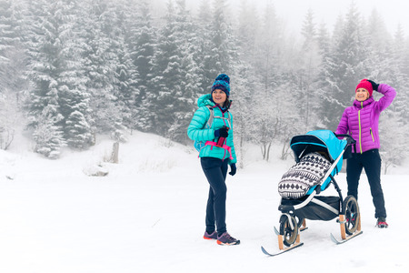Happy family in mountains. Mother with baby stroller enjoying motherhood in winter forest with female friend or partner. Hiking or power walking woman, sledge pram in woods, inspiring winter.の写真素材