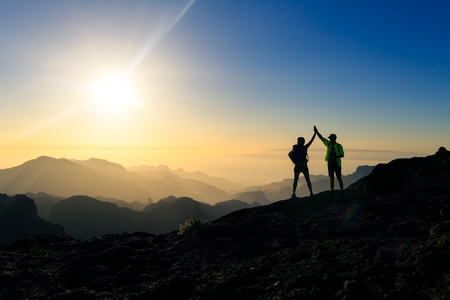 Couple hikers celebrating success in sunset mountains, accomplish with arms up outstretched. Young man and woman looking at beautiful inspirational landscape view, Gran Canaria Canary Islands.の写真素材