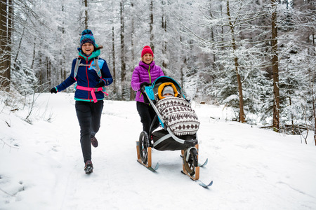 Mother with baby stroller enjoying winter forest with female friend or partner, family time. Hiking and running woman with sledge pram, teamwork concept. Beautiful winter inspirational mountains.の写真素材