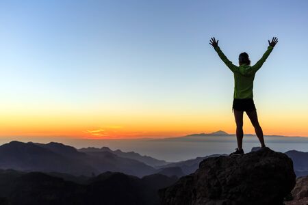 Woman successful hiking or climbing in mountains, motivation and inspiration in beautiful sunset landscape. Female hiker with arms up outstretched on mountain top looking at view.