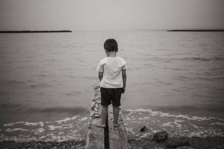 black and white photo of sad boy in white t-shirt and black pants on pier above water facing back to photographerの写真素材