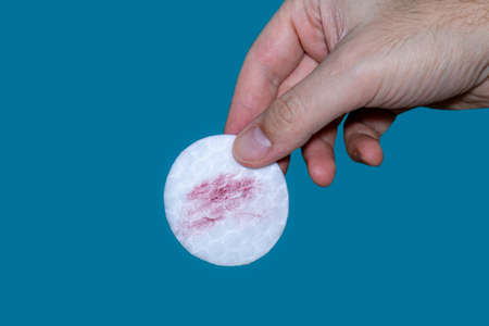 White cotton makeup pad with red lipstick traces held between fingers by Caucasian white male hand isolated on blue background 2020の写真素材