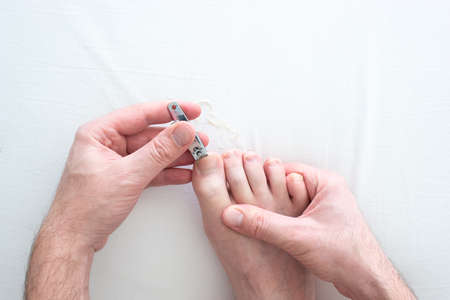 Caucasian male hand and foot cutting his toenails with a metal nail clipper on a white surface top view close up shot 2020の販売画像