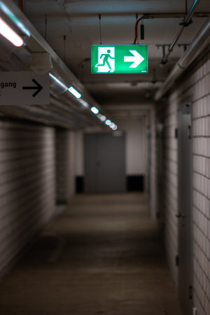 Illuminated green exit sign hanging from the wall in and underground poorly lit corridor 2020
