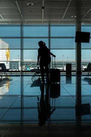 Caucasian young woman silhouette standing in an empty international airport terminal gate with a face mask in her hand next to her roller luggage 2020の写真素材
