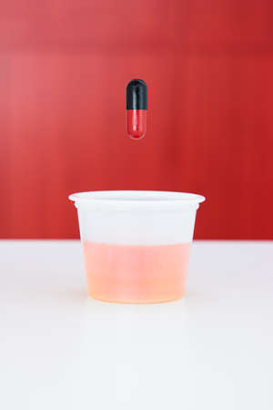Small transparent plastic medicine drinking cup with red and black medicament capsule above it shallow depth of field.の写真素材