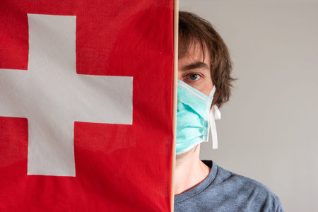 Caucasian young male with uncut hair wearing a green medical mask right side covered by Swiss national flag front view.の写真素材