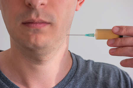 Unidentifiable Caucasian male holding a syringe with orange serum close to his neck shallow depth of field no eyes.の写真素材