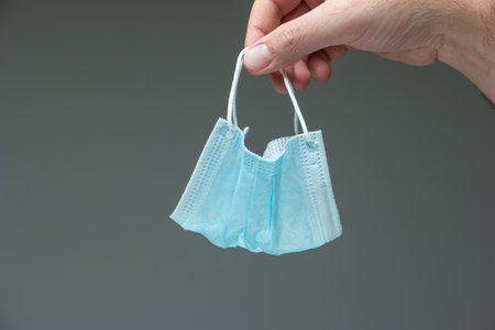 Caucasian male hand dangling a blue textile medical mask close up front view isolated on gray.の写真素材