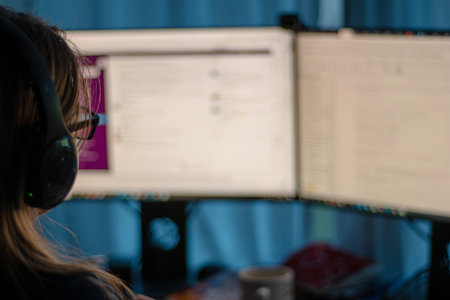 Unrecognizable woman with glasses working from home on two computer screen low key shot from behind.
