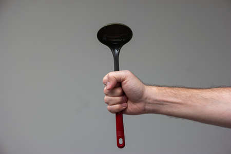 Caucasian male hand holding a black plastic kitchen ladle with red handle isolated on gray background.の写真素材