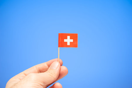 Small toothpick size Swiss national flag between fingers by Caucasian male hand isolated on blue...