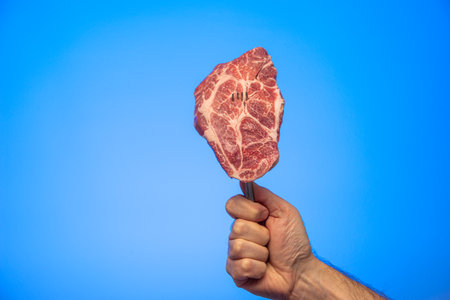 Slice of fresh raw marbled steak on a fork held in hand studio close up shot isolated on blue background.の写真素材