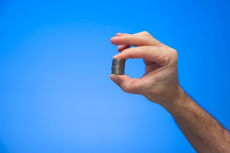 Caucasian male hand holding a stack of coins between fingers isolated on blue background studio shot.の写真素材