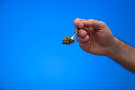 Caucasian male hand holding a spoon full of pickled capers isolated on blue background studio shot.の写真素材