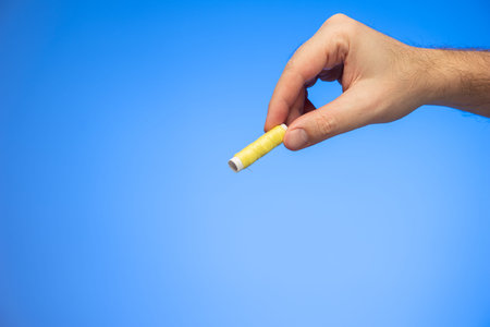 Small reel of yellow sewing thread held in hand by Caucasian male hand studio shot isolated on blue...